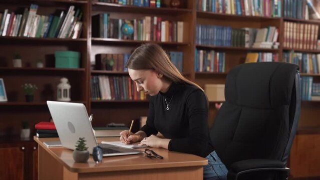 A girl is working in an office filled with books. She is sitting at a desk with a laptop and papers, focused on her work. Her facial expression indicates anxiety as something seems to be going wrong.