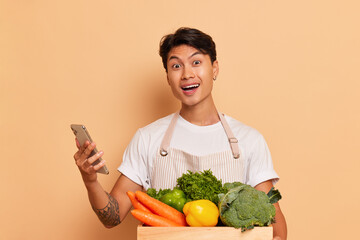 Young happy black-haired man stands against background, box full of fresh vegetables in front of...