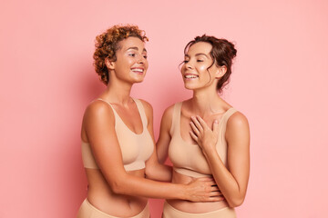 Two young smiling women posing half-nude against pink background wearing only underwear bras and panties, hugging and laughing, relationship concept, copy space - obrazy, fototapety, plakaty