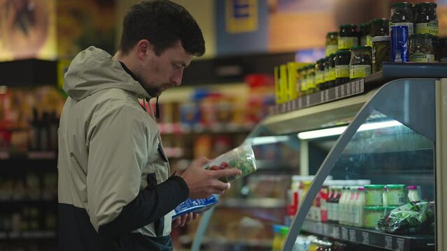 Man Chooses Salad While Holding Packet Of Milk In Hand At Supermarket In Jacket. Person Buys Groceries. Customer Picks Prepared Meal Packed Salad In Store. Healthy Eating. Guy Buys Box Of Lettuce. 