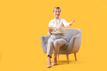 Fototapeta na wymiar Mature psychologist with tablet computer sitting in armchair on yellow background