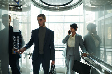 Young businessman pressing button in elevator while standing against female colleague with mobile phone by ear talking to client