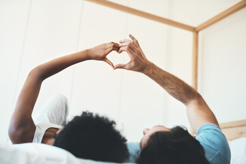 Interracial couple, hands and heart emoji in bed for morning, love or care relaxing together at home. Man and woman lying in bedroom with touching hand for loving symbol, sign or gesture at the house
