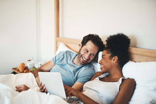 Happy couple, tablet and laughing on bed for joke, funny entertainment or online streaming at home. Interracial man and woman person on technology in relax for morning or social media in the bedroom