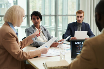 Blond female boss with financial document explaining something to group of young intercultural subordinates during discussion of data