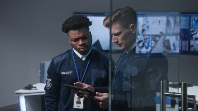 Male officer uses tablet computer and talks to African American colleague. Multi ethnic workers in security control center. Big digital screens with surveillance CCTV cameras footage on background.