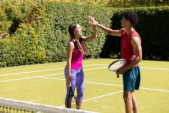 Happy diverse couple holding rackets and high fiving on sunny outdoor tennis court - Powered by Adobe