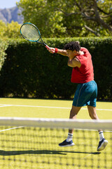 Fototapeta na wymiar Biracial man returning ball with racket on sunny outdoor tennis court