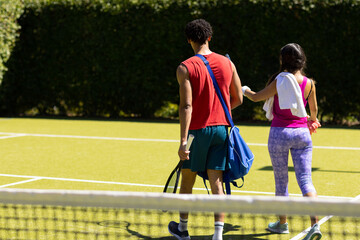 Obraz premium Rear view of diverse couple with bags and rackets walking on sunny outdoor tennis court, copy space