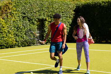 Obraz premium Happy diverse couple with bags and rackets walking on sunny outdoor tennis court