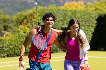 Obraz premium Happy diverse couple with bags and rackets embracing on sunny outdoor tennis court