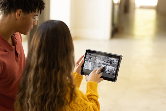 Diverse couple at home looking at tablet with security camera views on screen