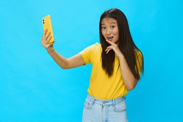 Happy Asian woman holding her phone and looking at the screen taking selfies with a yellow case on a blue background in a yellow T-shirt smiling with teeth 