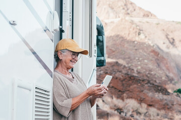 Happy senior woman with eyeglasses standing outside a camper van motor home using mobile phone...