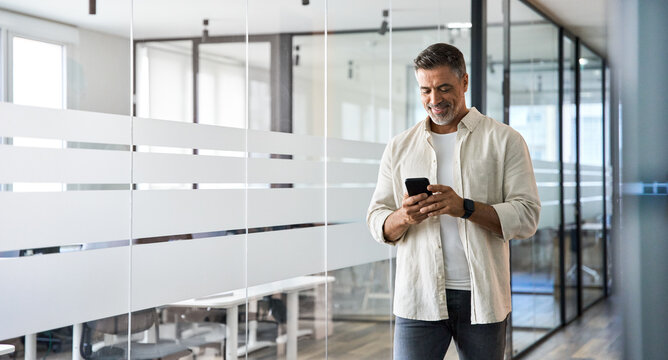 Mature Latin Or Indian Businessman Walking With Smartphone In Office. Middle Aged Manager Using Cell Phone Mobile App. Digital Technology Application And Solutions For Business Success Development.