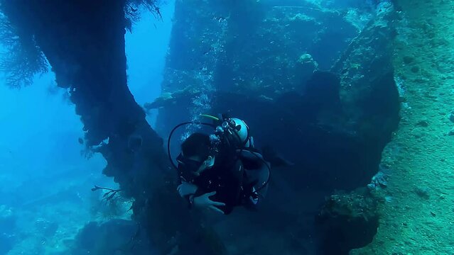 A man is scuba diving underwater inside a shipwreck. He swims up and past the camera. The water is clear. 
