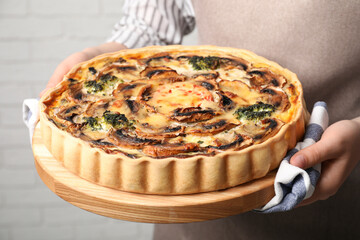 Woman holding delicious homemade quiche with mushrooms near white brick wall, closeup