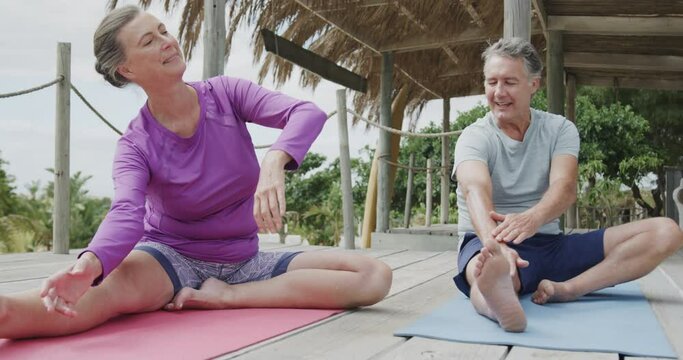 Happy senior caucasian couple practicing yoga sitting on beach sun deck, in slow motion