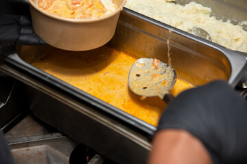 A view of a worker scooping sauce into a paper container, in a restaurant kitchen setting.