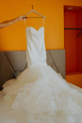 Arm holding a white bridal gown over a bed in a room with orange