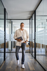 Vertical portrait Latin Hispanic mature adult professional business man, smiling Indian senior businessman CEO walking and holding digital tablet with online application inside modern company office.