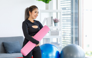 Obraz na płótnie Canvas Asian beautiful healthy fit slim female sporty athlete model in black sports bra and leggings standing smiling holding pink yoga mat ready to workout exercise in living room at home with rubber ball