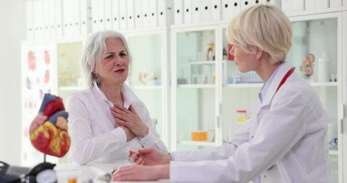 Elderly woman complains of acute chest pain to family doctor sitting next to heart model. Lady specialist listens to client complaints analyzing causes slow motion