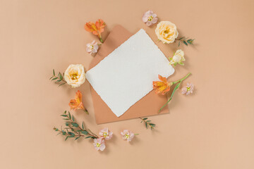 Directly above shot of empty handmade cotton paper, envelope, flower buds, green twigs  on beige background. Branding, wedding, birthday mockup.
