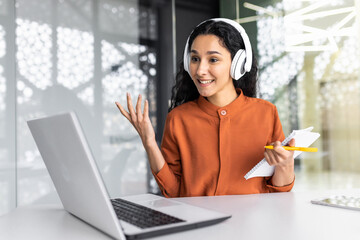 Young beautiful woman listening to online course in headphones and talking to mentor on video call using laptop, Latin American self-improvement learns at workplace inside office.