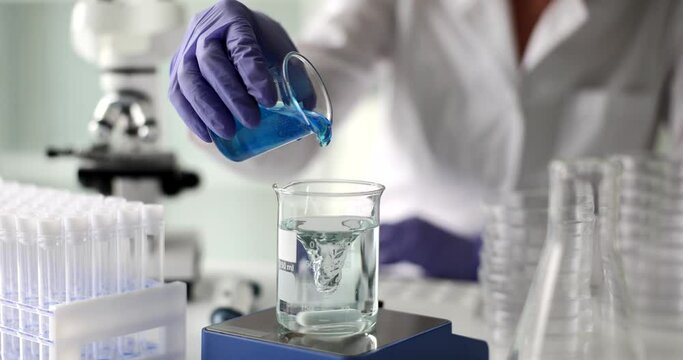 Scientist pours dense blue reagent into glass with swirling clean water. Woman in rubber gloves conducts scientific experiment in modern laboratory slow motion