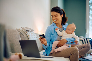 Happy mother shopping online with credit card while being with her baby daughter at home.