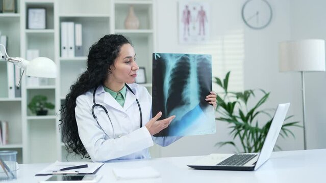 Female doctor talking on a video call using a laptop while holding an x-ray in a modern hospital clinic. Medical worker physician consults a patient sitting at a desk at a workplace in an office