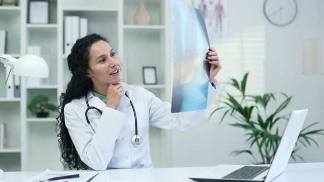 Confident female doctor in white coat examining x-ray picture while sitting at workplace in office in hospital clinic. Brunette medical worker physician is carefully studying a picture of the lungs