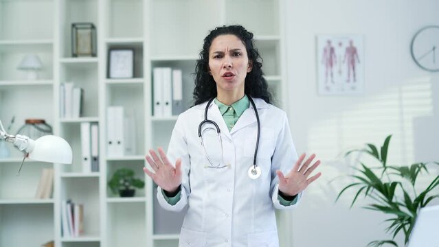 Female doctor in white coat showing crossed arms, X sign, shaking head no while standing in modern hospital clinic. Medical worker physician demonstrates a gesture of prohibition, denial, disagreement