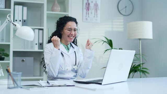 Happy excited female doctor in white coat read great news on laptop while sitting at desk at workplace in modern hospital clinic. Joyful surprised medical worker physician celebrates success in office