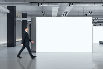 Marketing concept with businessman walking by blank white partition with space for advertising poster or picture frame in loft style gallery hall with concrete floor and grey wall background, mockup - obrazy, fototapety, plakaty