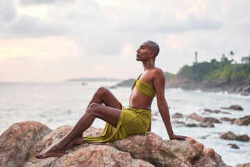 Gender fluid black person poses gracefully sitting on rocks in ocean at sunset. Androgynous slim ethnic fashion model in tropical maxi dress on top of rocky hill above storm at dusk. Pride LGBTQIA