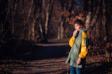 Young girl in yellow-green raincoat standing in autumn forest park. Eating food with strange face