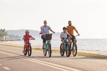 Fototapeta premium Two kids, with helmets on their heads, and smiling parents riding bikes on a family-friendly cycle route along a sea coastline, front view.