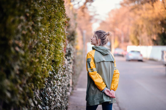 Young woman with funny ponytail in green raincoat stand near old wooden fence, yellow blooming tree