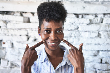 Obraz premium Radiant middle-aged African woman smiles in her living room with white brick wall and natural light.