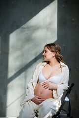 A stylish pregnant young woman in a white suit sits on a chair and touches her belly with a gentle smile. Awaiting birth. Care and motherhood. Love and tenderness.