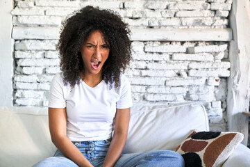 A young african american woman sitting on her sofa screaming very angry and aggressive.