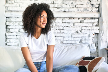 Fototapeta na wymiar A young african american woman sitting on her sofa looks aside smiling, cheerful and pleasant.