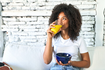 African-American woman enjoys nourishing breakfast of oatmeal and orange juice on the sofa,...