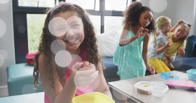Animation of white bokeh light spots over happy diverse schoolgirls eating packed lunch and talking