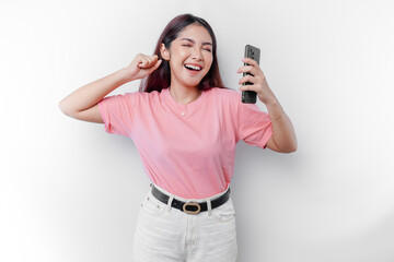 A young Asian woman with a happy successful expression wearing pink t-shirt and holding smartphone isolated by white background