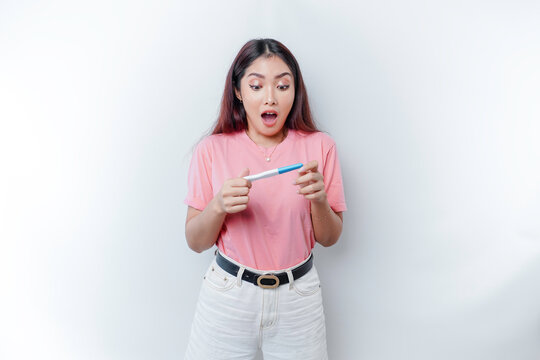 Unhappy woman wearing pink t-shirt looking at pregnancy test alone, confused young female shocked by result, bad news, covering open mouth with hand, unwanted pregnancy concept