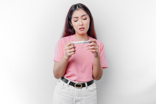 Sad woman wearing pink t-shirt looking at pregnancy test alone, shocked by result, bad news, covering open mouth with hand, unwanted pregnancy concept