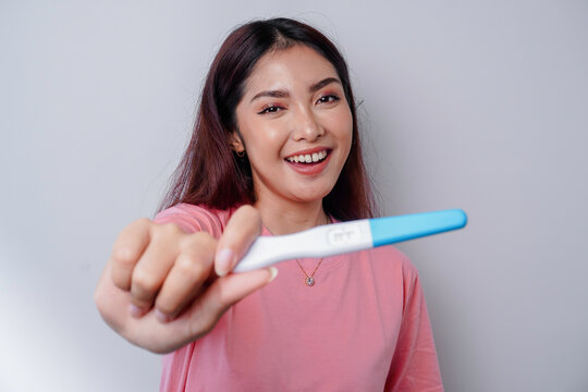 Happy young woman wearing pink t-shirt showing her pregnancy test, isolated on white background, pregnancy concept